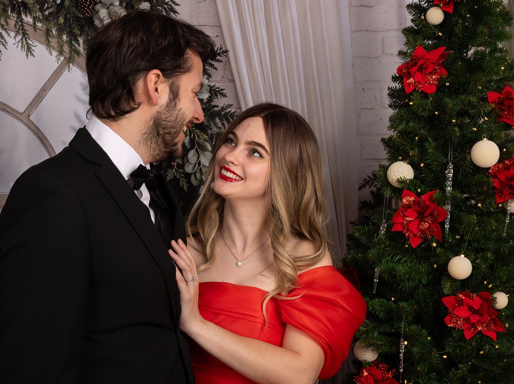 A smiling woman in a red off-shoulder dress gazes at a man in a black tuxedo and bow tie beside a decorated Christmas tree.