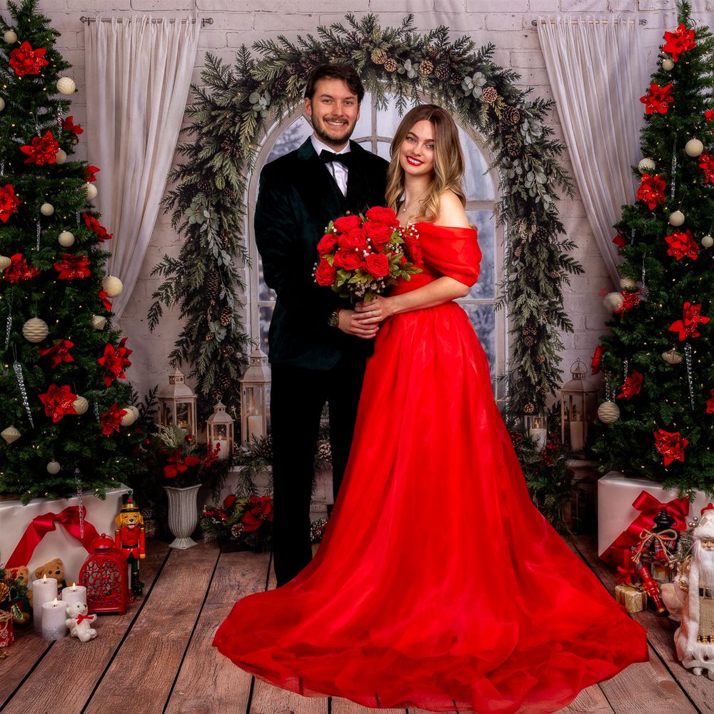 Couple in a red off-shoulder gown and black tuxedo holding a red rose bouquet in a Christmas-themed studio with decorated trees.