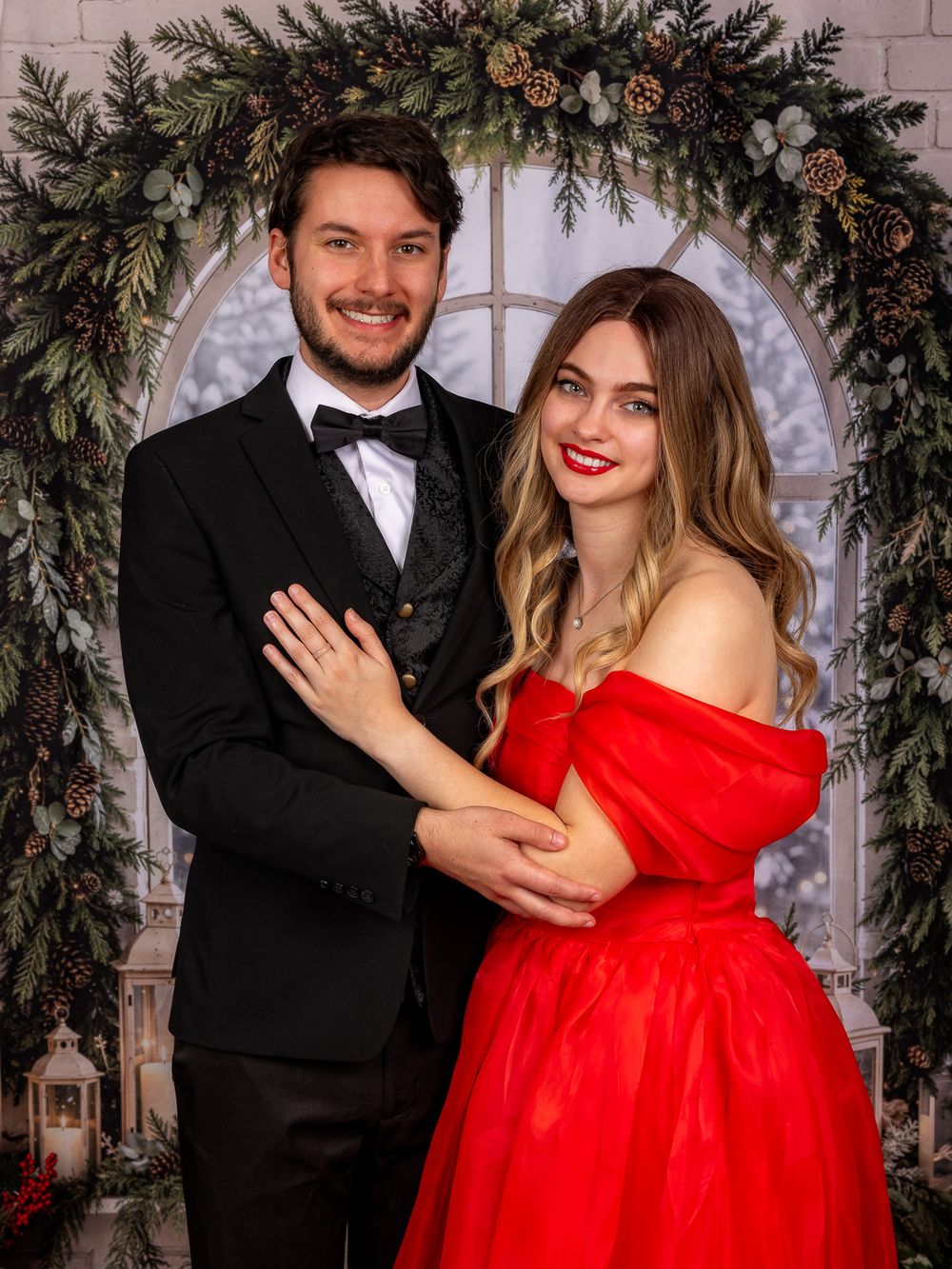Smiling couple in formal wear, a man in a black tuxedo and a woman in a red off-shoulder gown, posing before a Christmas greenery arch.