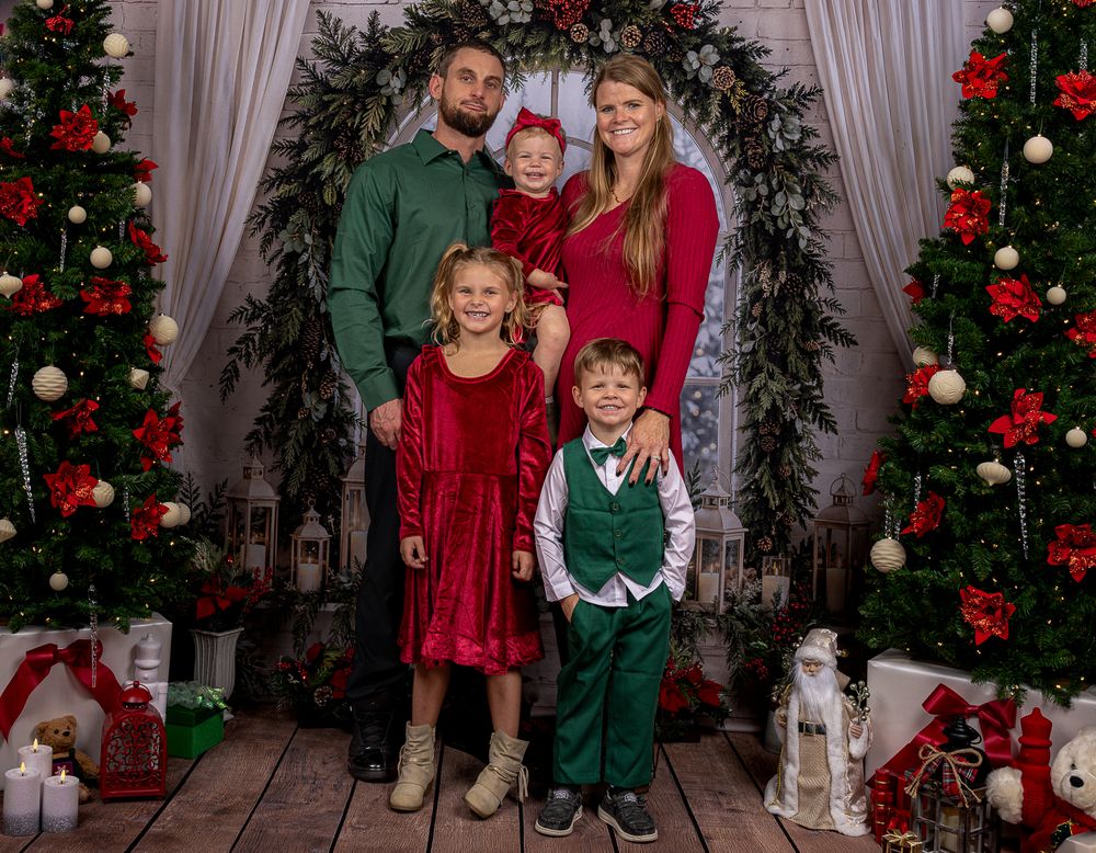 Family of five in red and green outfits poses for a Christmas portrait with decorated trees, a greenery wreath and gifts on a wooden floor.