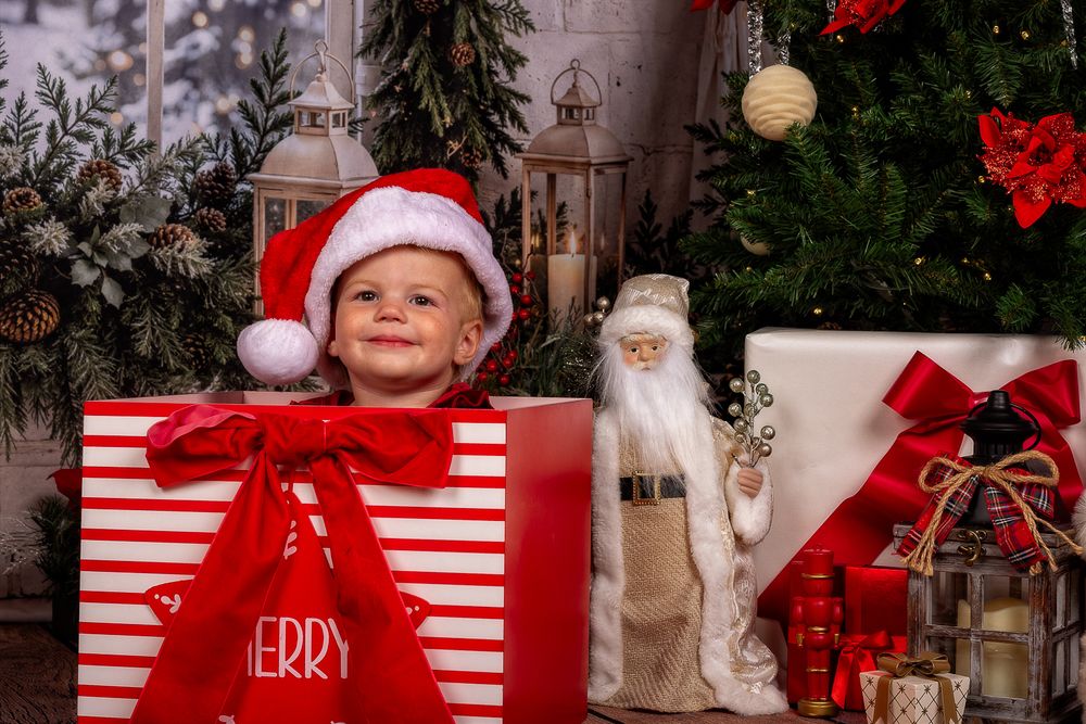Smiling child in a Santa hat peeks over a striped Christmas gift box with red bow beside a Santa figurine and decorated tree.