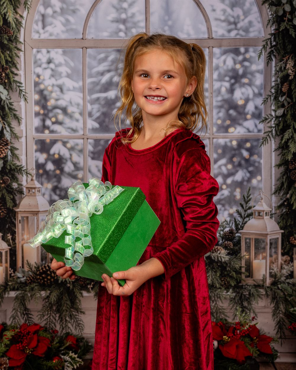 Smiling girl in a red velvet dress holds a green gift box with a sparkly bow in a festive Christmas photo studio setting.