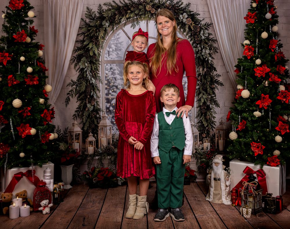 Smiling mother and two children in red and green outfits pose in a festive Christmas photo studio with decorated trees and gifts.