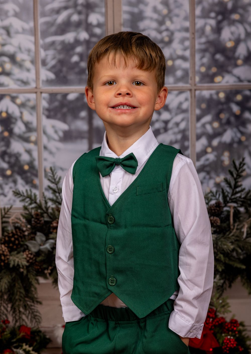 Smiling young boy in a green vest, white shirt and green bow tie poses in front of a snowy window with holiday evergreen garland.