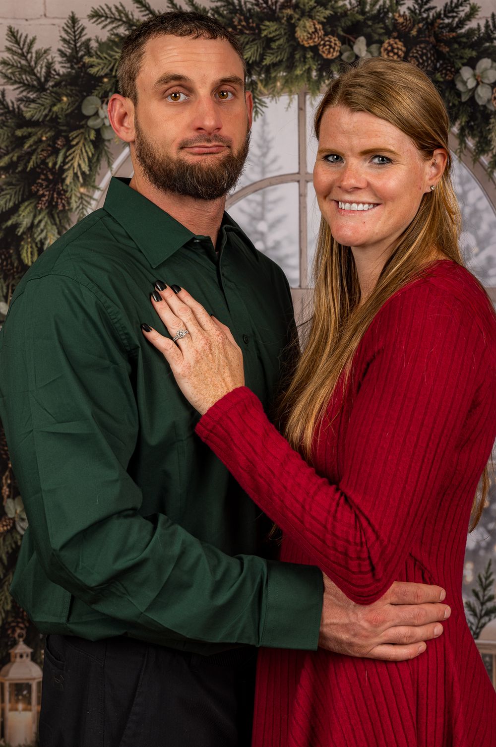 Smiling couple in a green button-up shirt and red knit dress posing together in front of a festive holiday backdrop with evergreen garland.