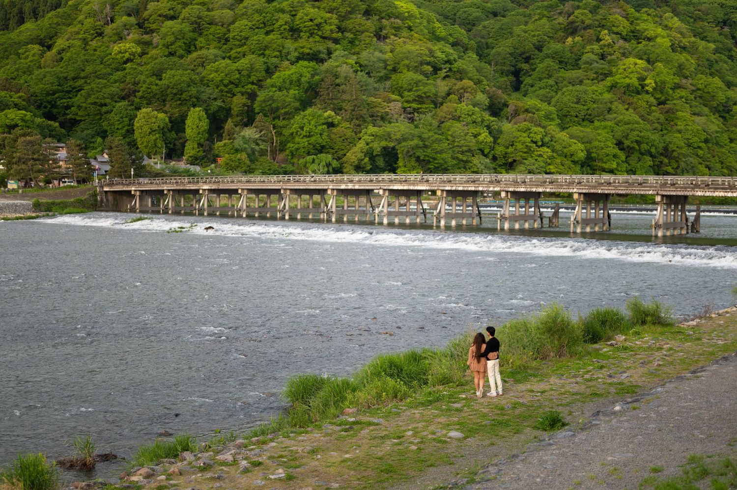 Couple stands on a grassy riverbank beside a wide, flowing river with a long wooden bridge and lush green forested hills in the background.