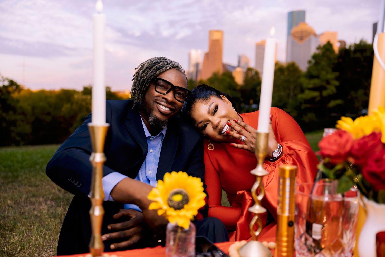 Smiling couple sits close at a candlelit outdoor picnic table with sunflowers and roses, a city skyline glowing behind them at sunset.
