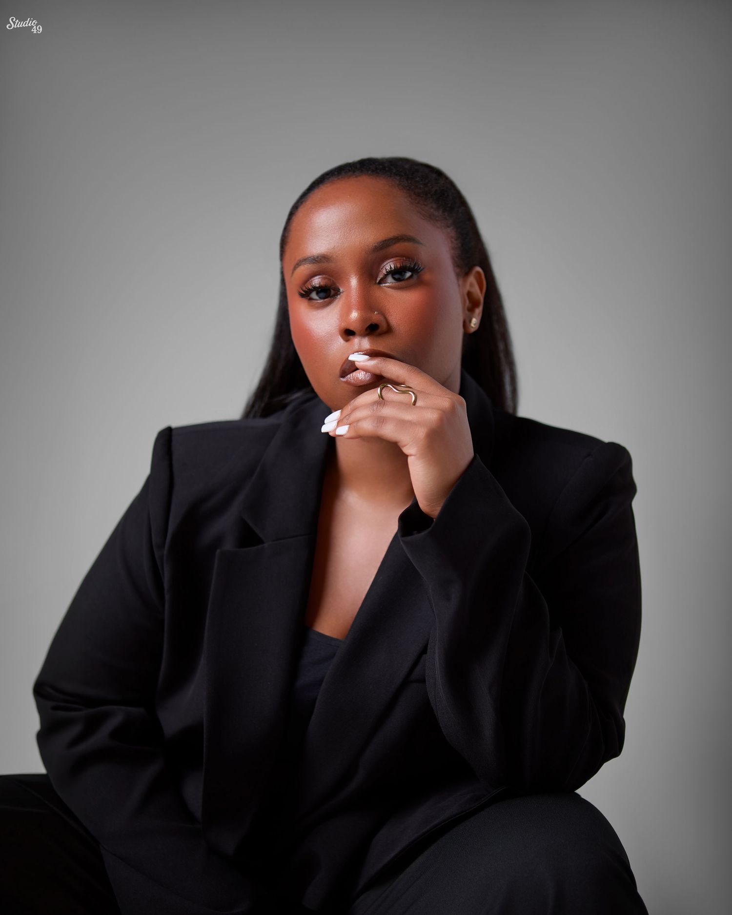 A young Black woman in a black blazer poses confidently with hands clasped near her chin against a gray background.