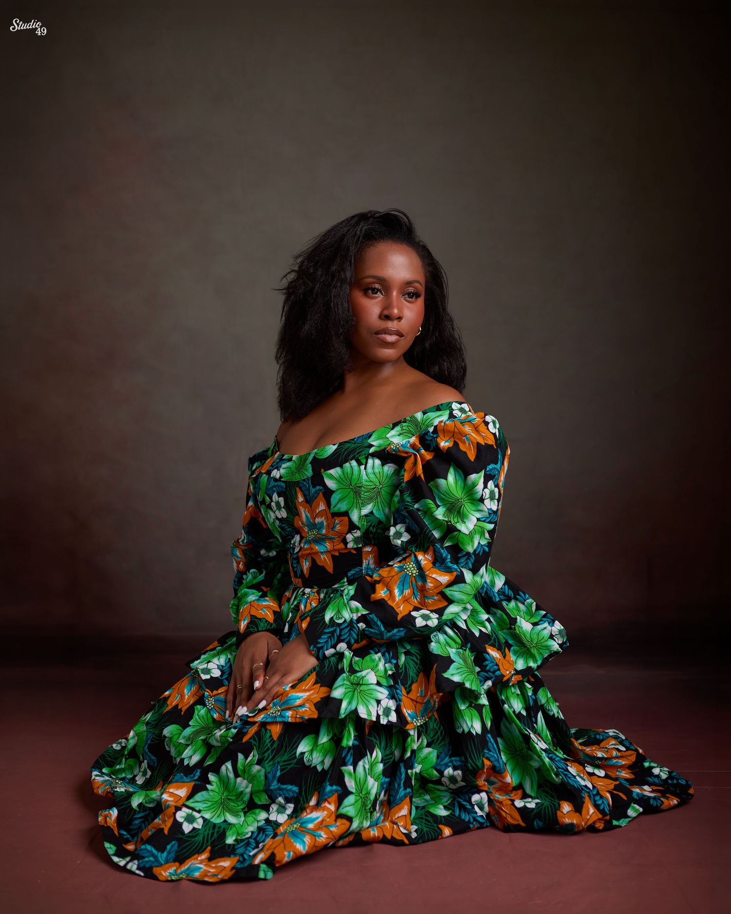 Woman in vibrant green floral off-shoulder dress seated gracefully against a dark studio background.