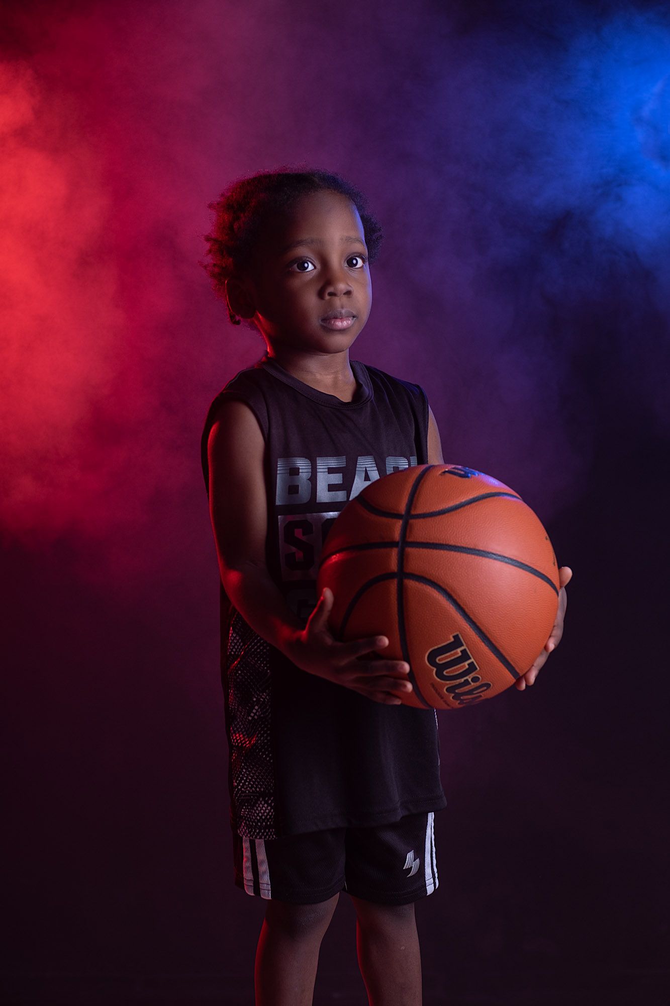 Young boy in basketball uniform holding a Wilson basketball against dramatic red and blue smoke background.