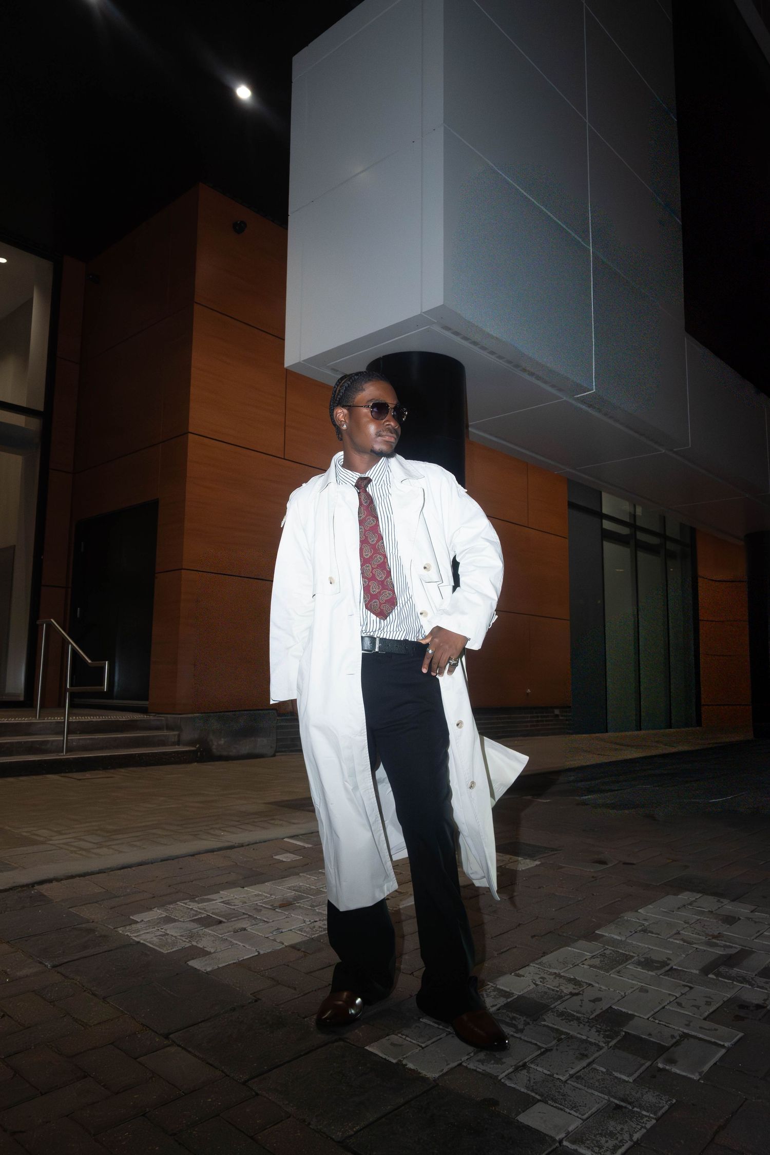 Man in white trench coat and red tie poses confidently outside modern building at night wearing sunglasses.