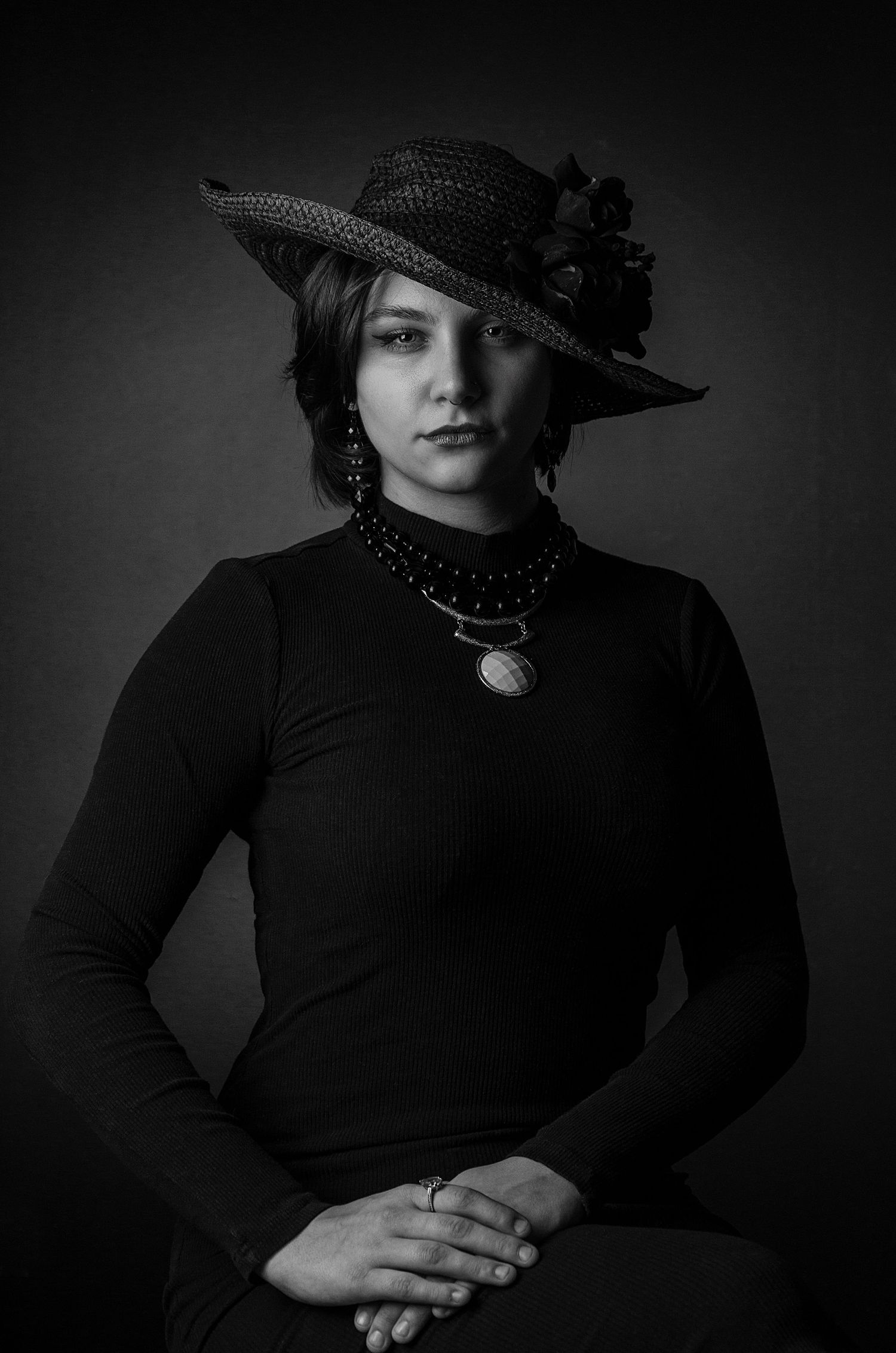 Black and white portrait of a woman in a wide-brim hat, black turtleneck, and layered necklaces, posing elegantly.
