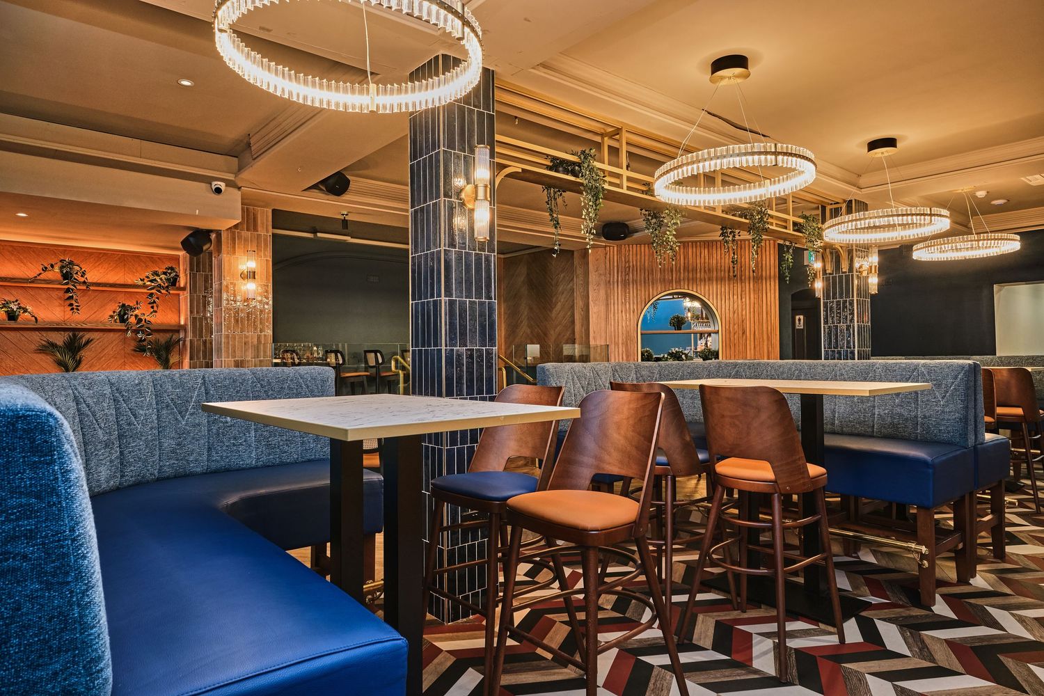 Sophisticated restaurant interior with blue velvet booths, circular chandeliers, and herringbone patterned flooring.