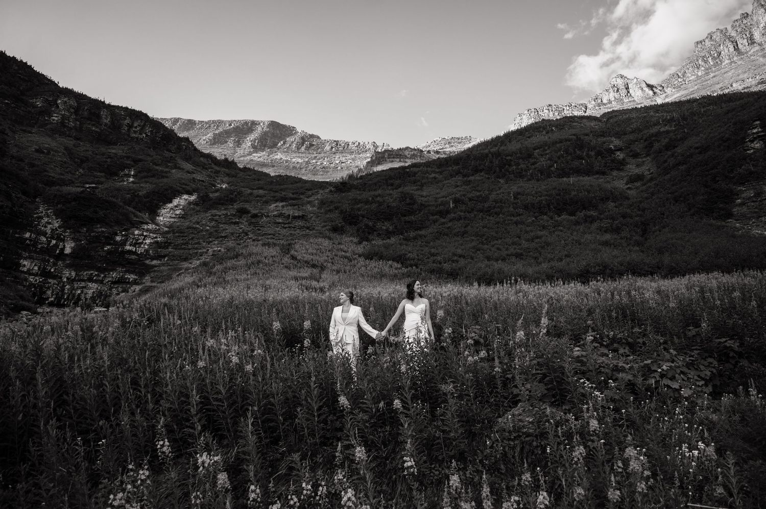 Black and white photo of a couple holding hands in a wildflower meadow with dramatic mountain peaks in the background.