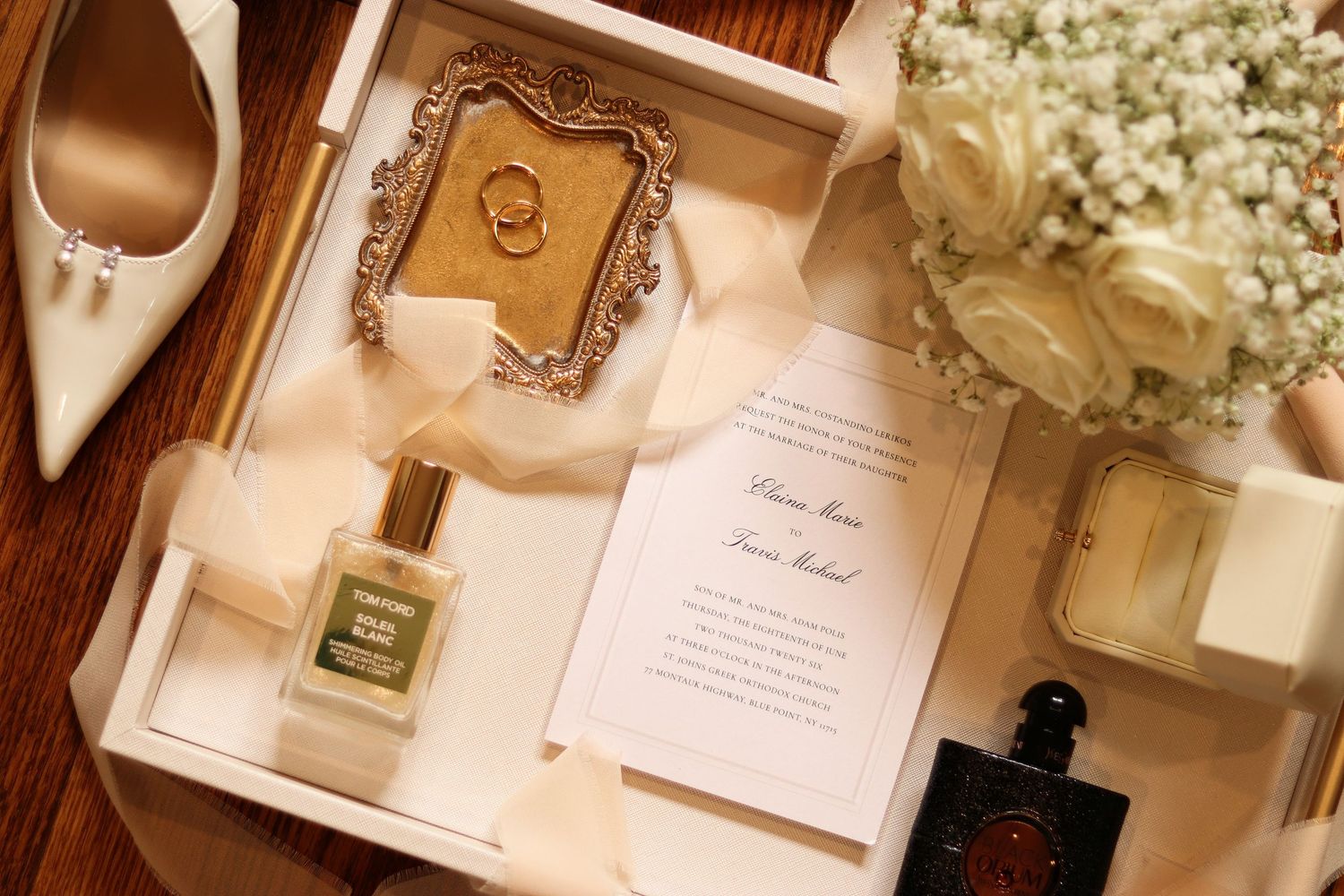 Elegant flat lay of a wedding invitation, gold ring frame, perfume bottles, white rose bouquet and bridal heels on a rustic wooden table.