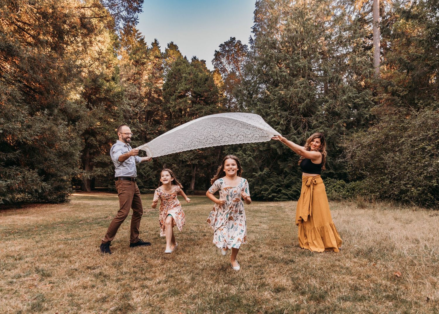Two parents hold a sheer blanket overhead while two young girls in dresses run beneath it on a grassy lawn surrounded by tall trees.