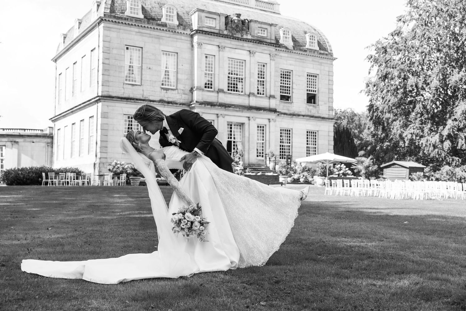 Black and white photo of bride and groom sharing a romantic dip kiss on lawn before grand estate wedding venue.