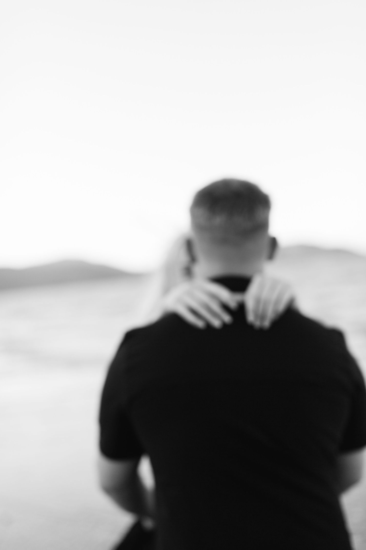 Black and white blurred photo of a couple embracing outdoors, woman's hands resting on man's neck.