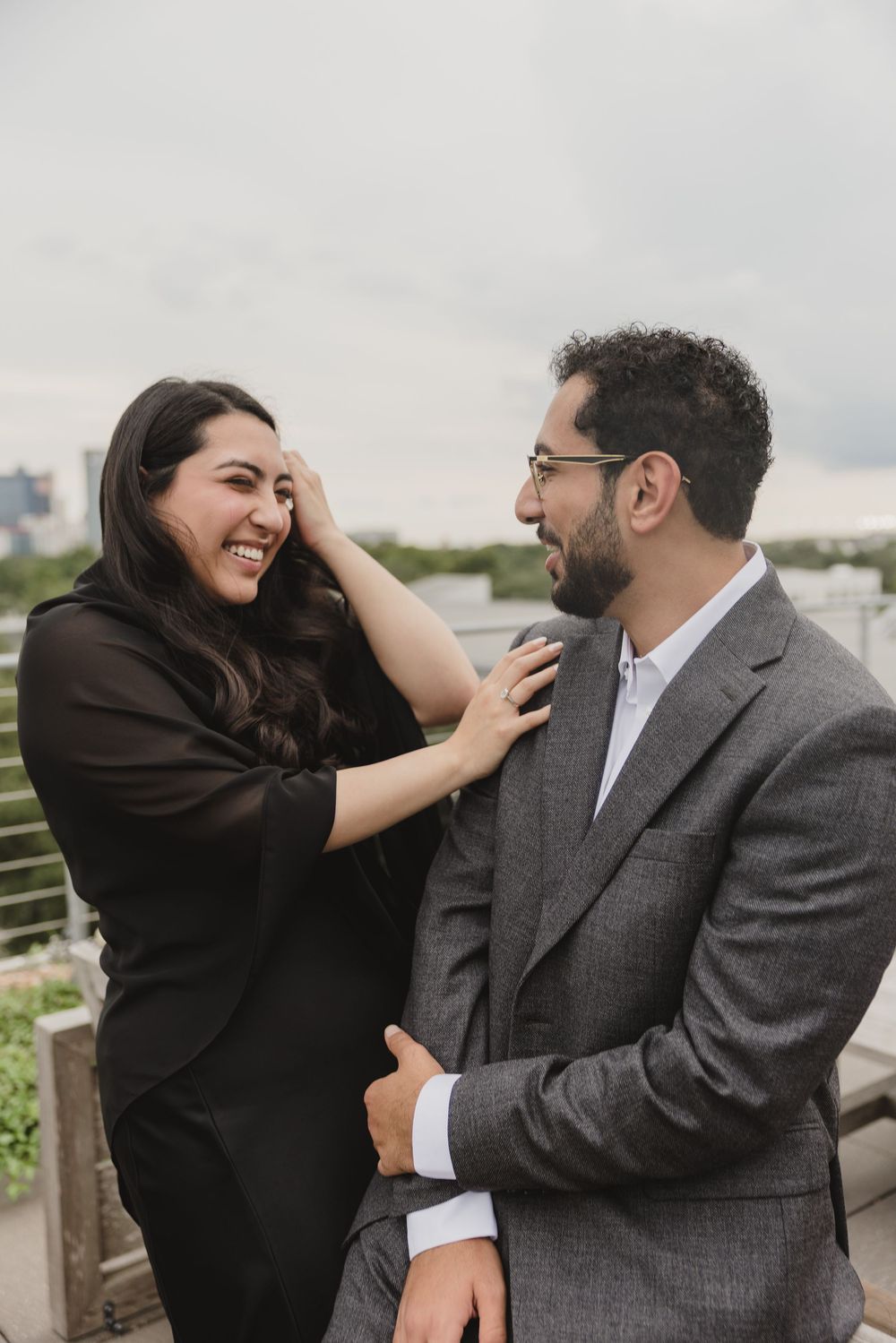 A laughing couple in formal attire share a joyful moment on a rooftop, the woman touching the mans face affectionately.