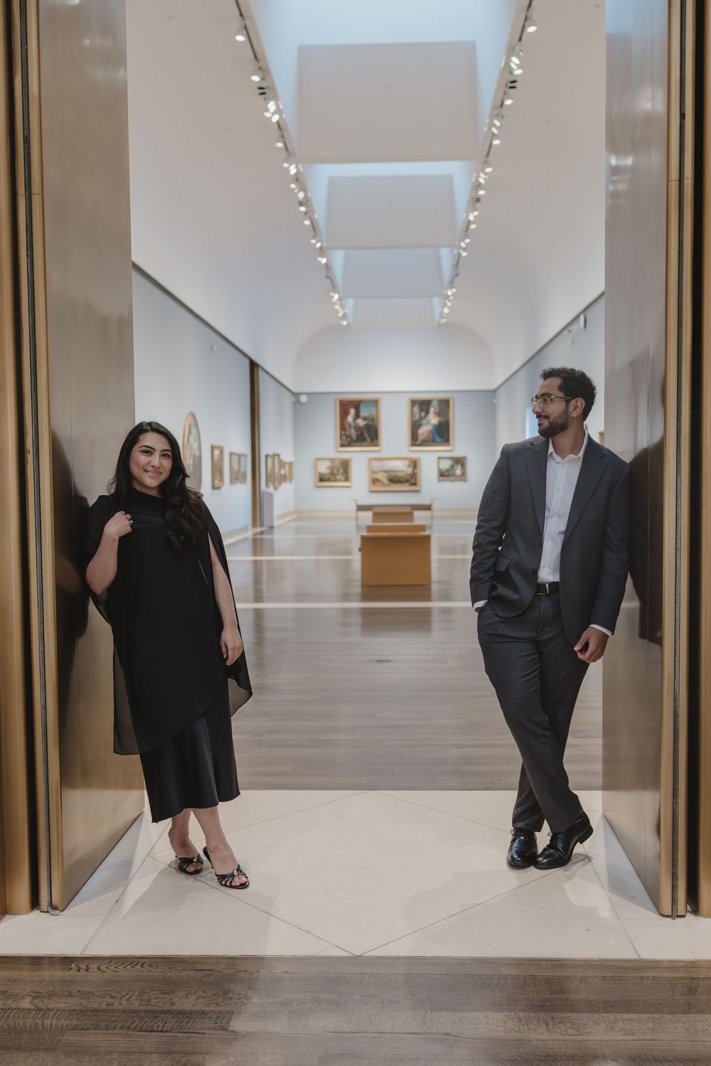 A man and woman in formal attire stand at the entrance of an elegant art gallery hallway with paintings.