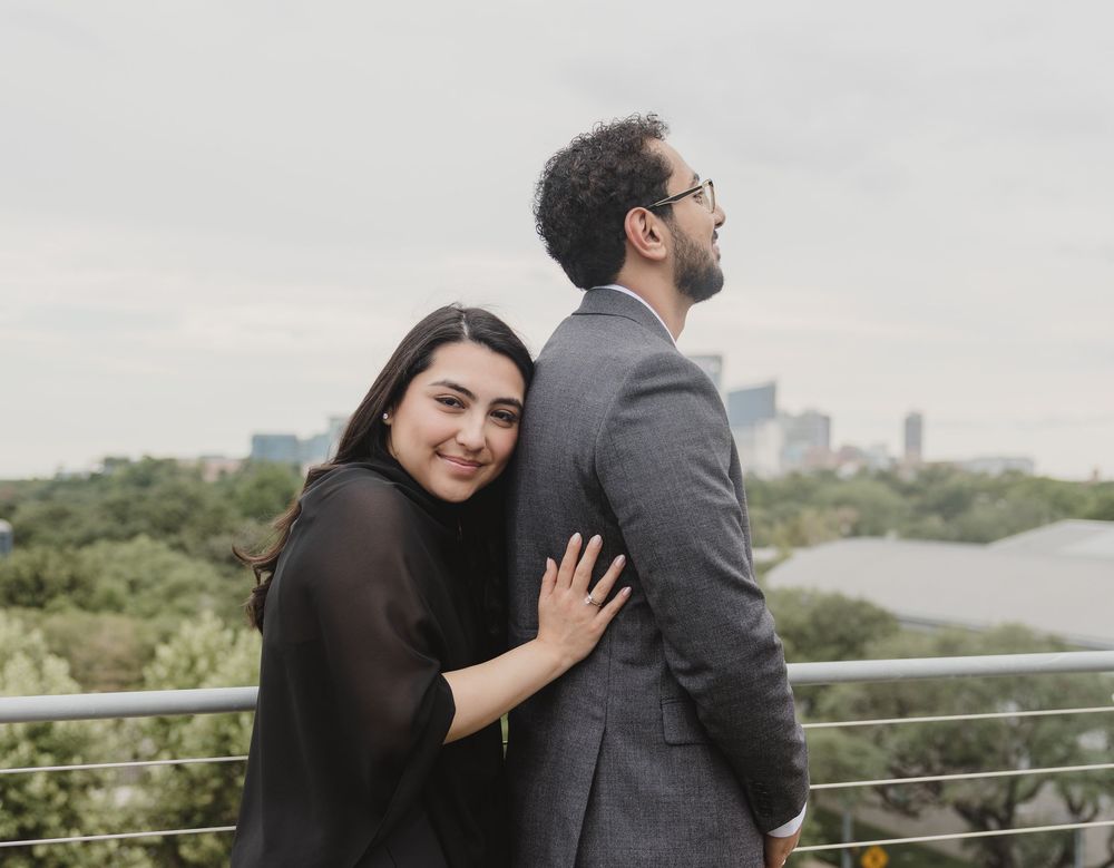 A woman in black hugs a man in a gray suit on a rooftop with a city skyline and green trees behind them.