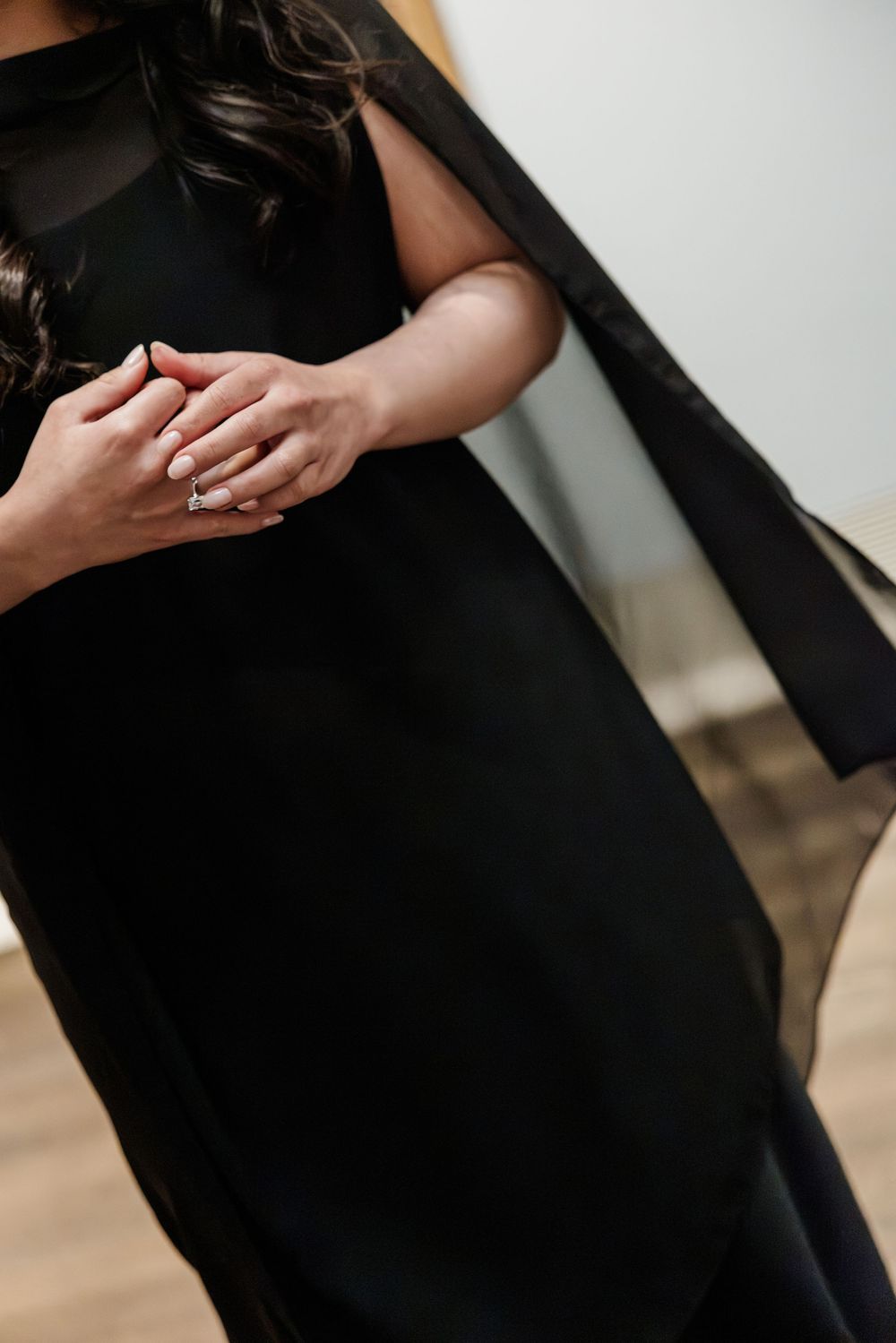 Woman in elegant black cape dress clasping hands, showcasing a diamond ring, standing on hardwood floor.