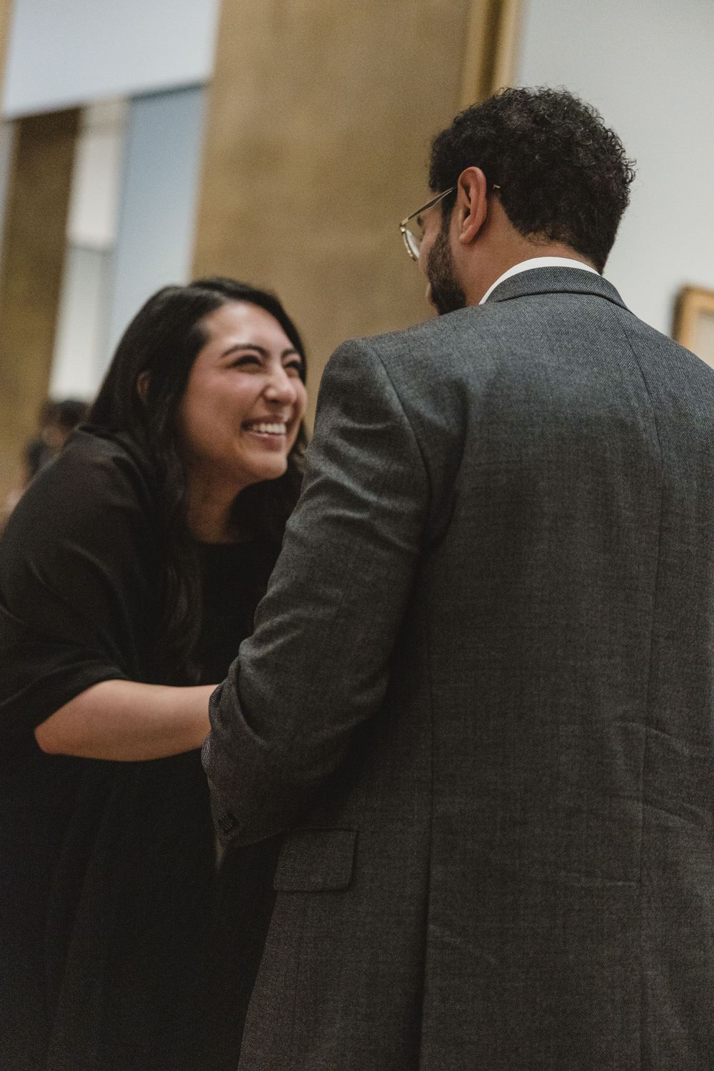 A smiling woman in black engages warmly with a man in a gray suit at an indoor social gathering.