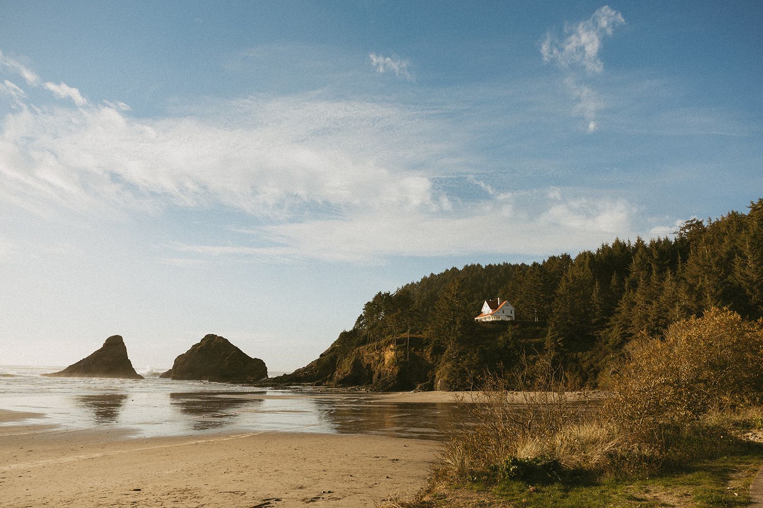 Scenic coastal view through lush green trees revealing a sandy beach and ocean inlet surrounded by forested cliffs.