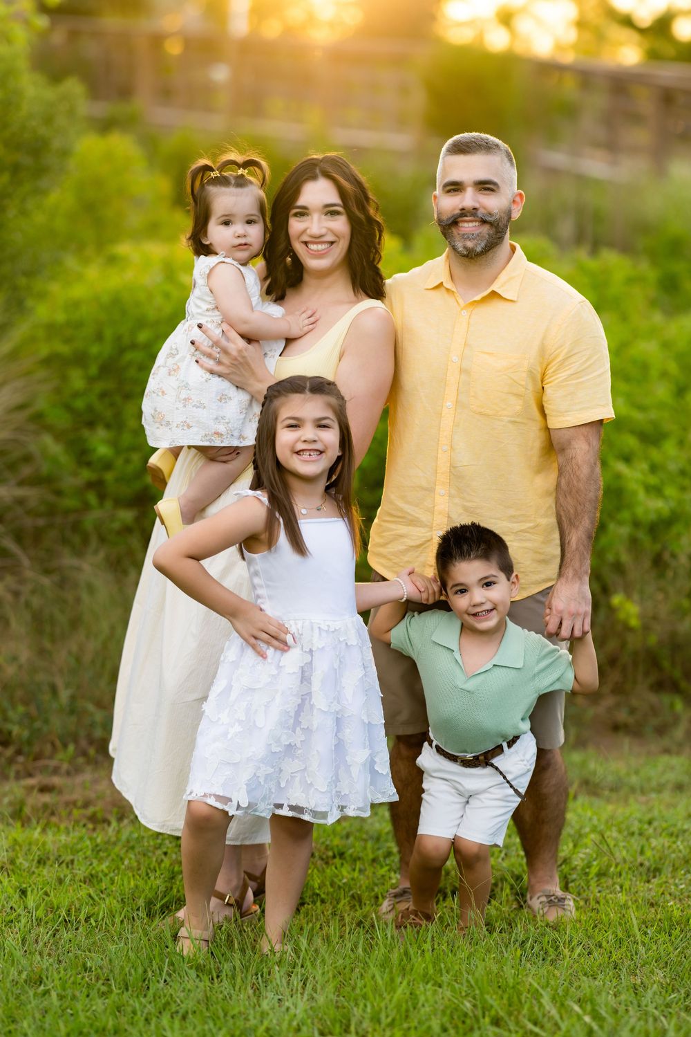 Happy family of five posing outdoors at golden hour on green grass, parents with three young children smiling.