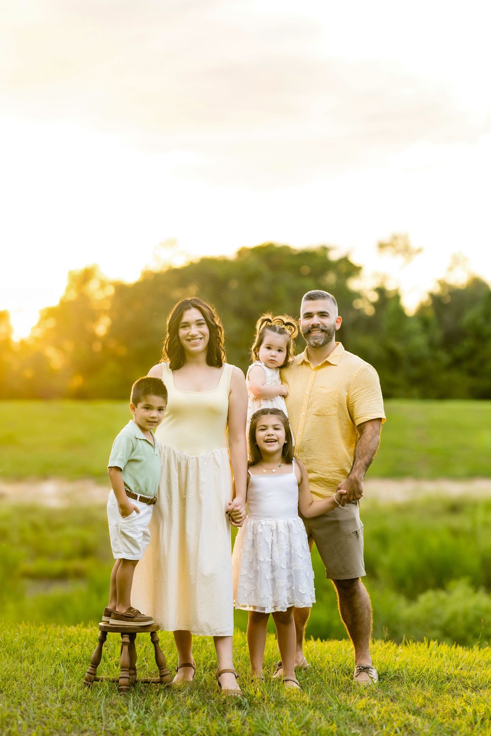 Happy family of five posing outdoors at golden hour in a green field, wearing coordinated yellow and white outfits.