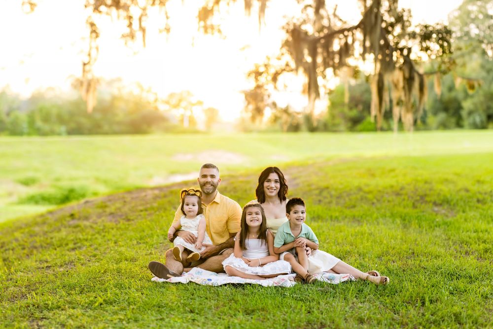 Family of five sitting on grass at golden hour under Spanish moss-covered oak trees in a lush green park.