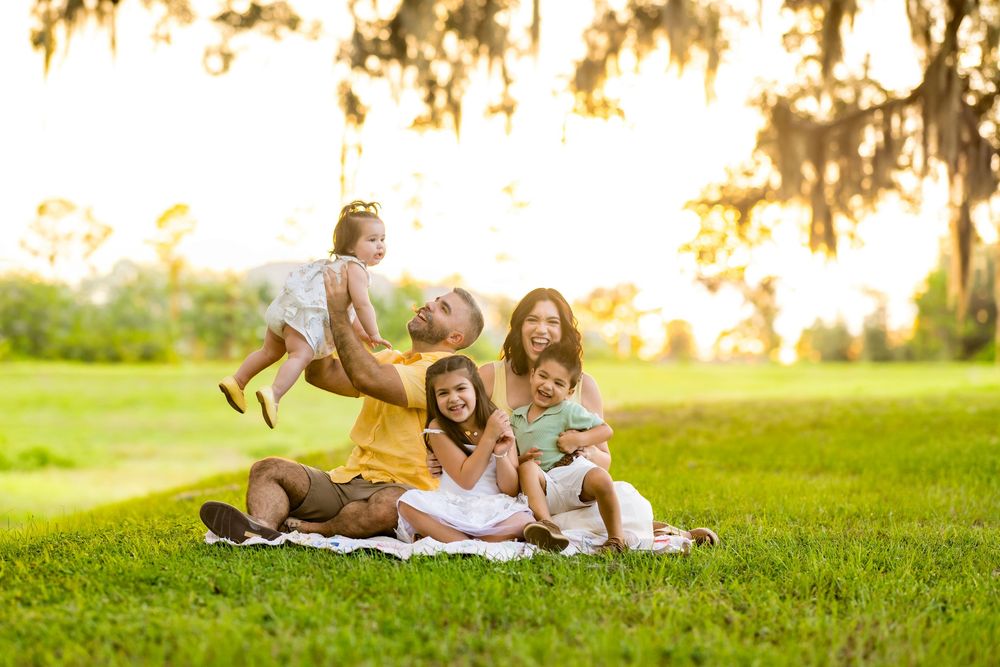 Happy family of five sitting on grass in a sunny park, parents lifting toddler in the air.