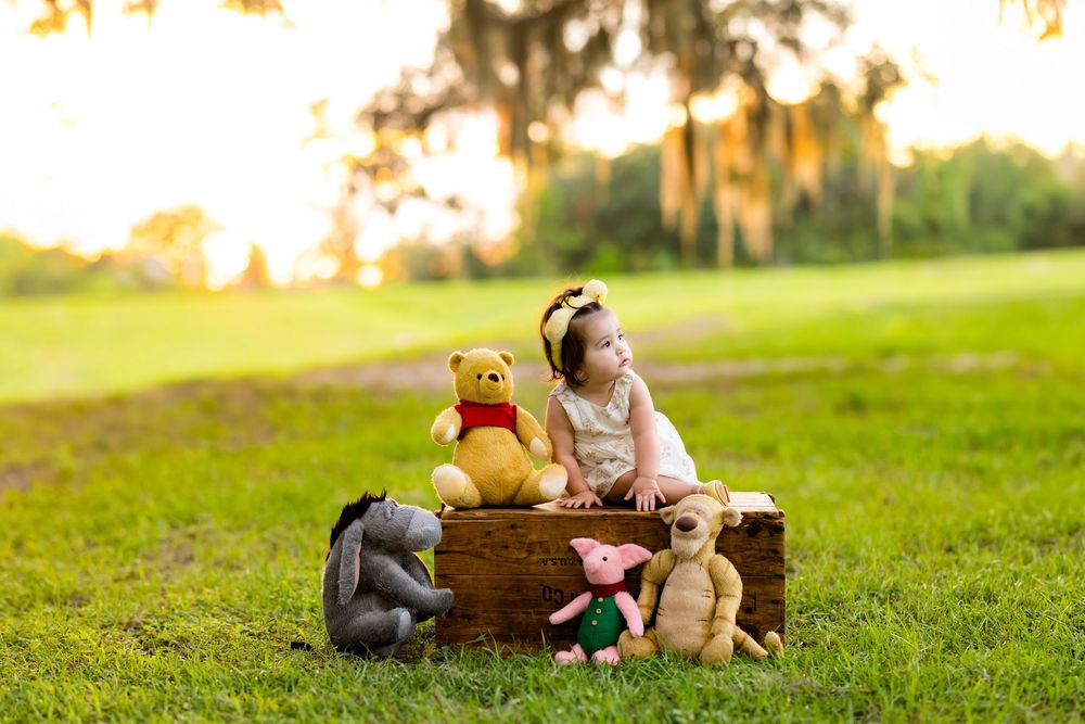 A toddler girl sits in a wooden crate outdoors with Winnie the Pooh stuffed animals at golden hour.