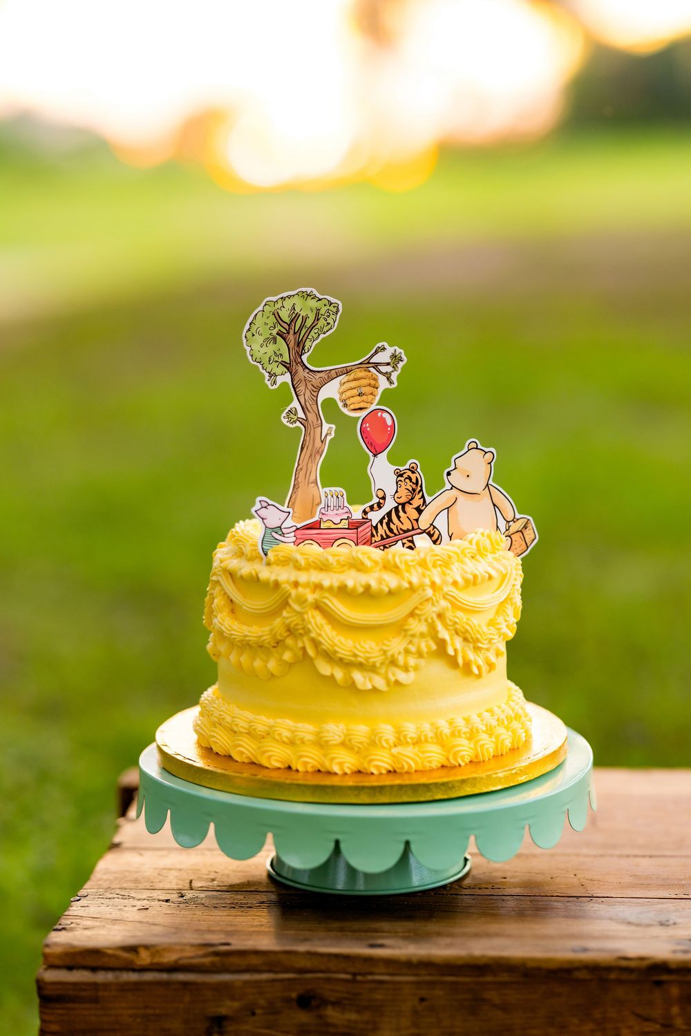 Yellow Winnie the Pooh themed birthday cake with character toppers on a mint cake stand outdoors.