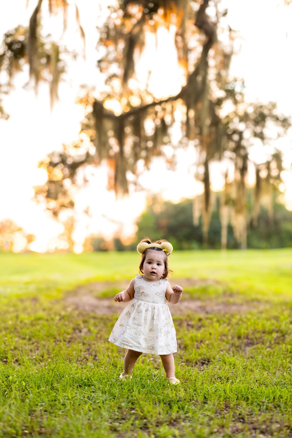 Toddler girl in white dress and floral headband standing on grass under Spanish moss trees at sunset.