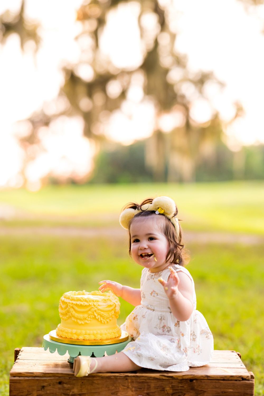 A joyful baby girl in a white dress smashes a yellow birthday cake outdoors during a first birthday cake smash photo session.