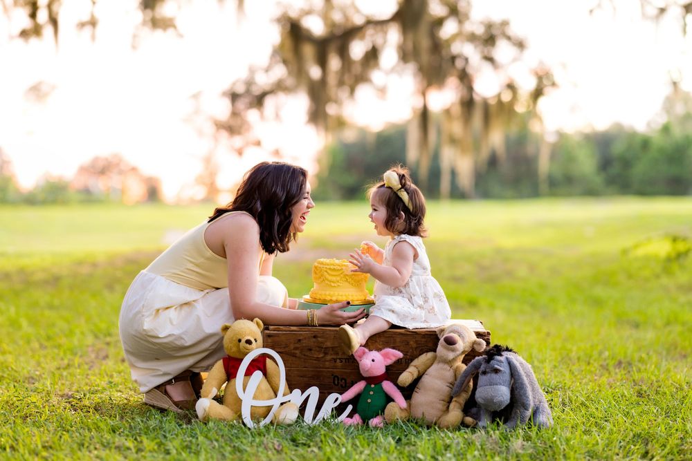 Mother and toddler girl celebrate first birthday outdoors with Winnie the Pooh stuffed animals and a yellow cake on green grass.