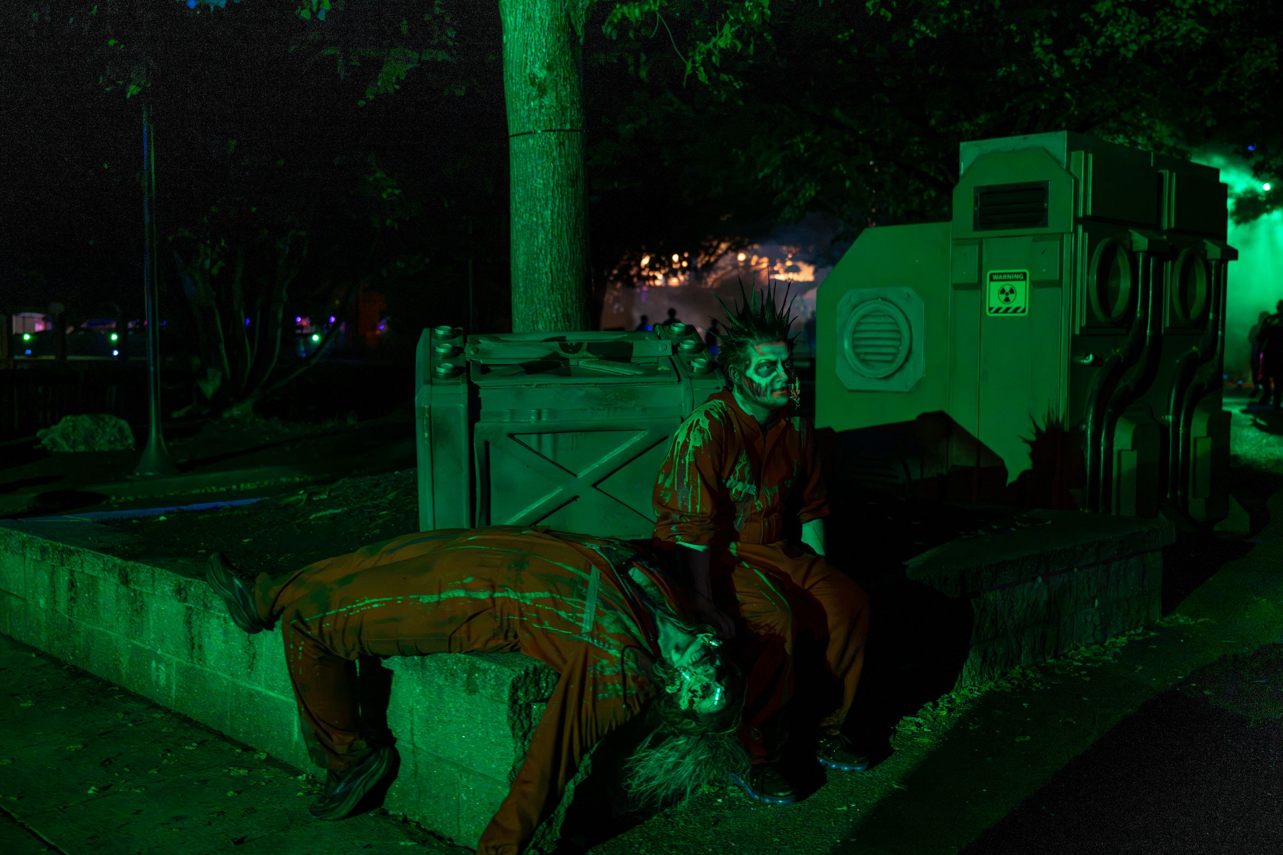 Spooky Halloween scene with a clown figure sitting under green lights near rustic props and a military vehicle at night.