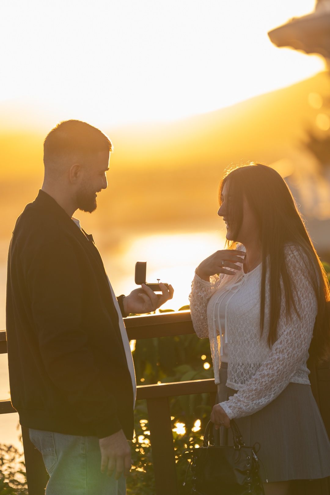 Man proposing to woman with ring box at golden sunset outdoors.