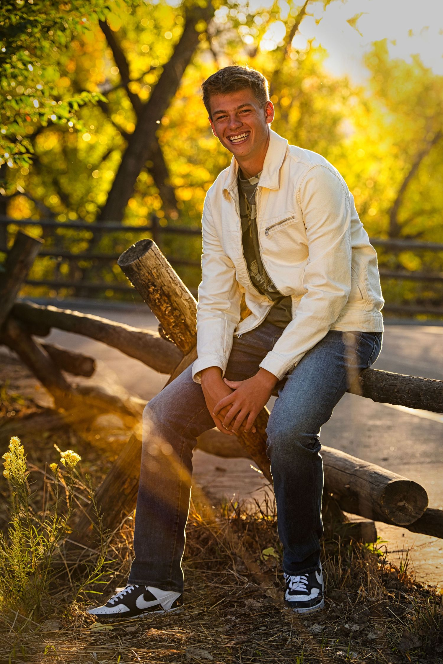 Young man in white jacket and jeans sits on rustic log fence outdoors in golden autumn sunlight.