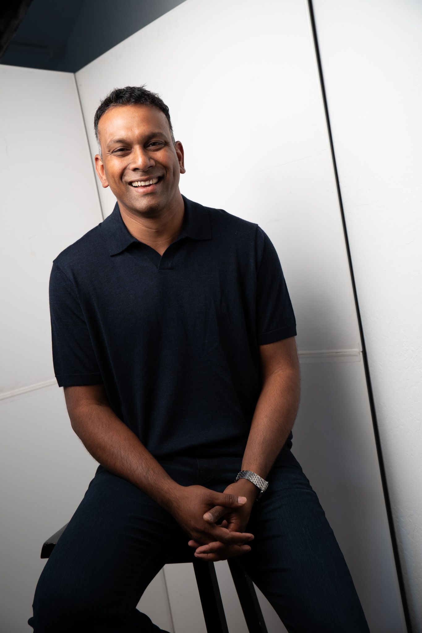 Smiling man in navy polo shirt sitting on stool against white background, hands clasped, wearing a silver watch.