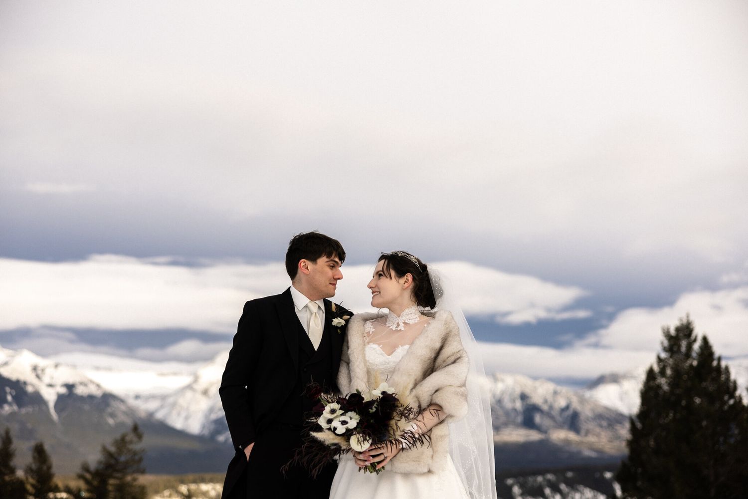 Bride and groom on the snowy Fairmont Banff Springs terrace with snow-capped mountains and dramatic clouds