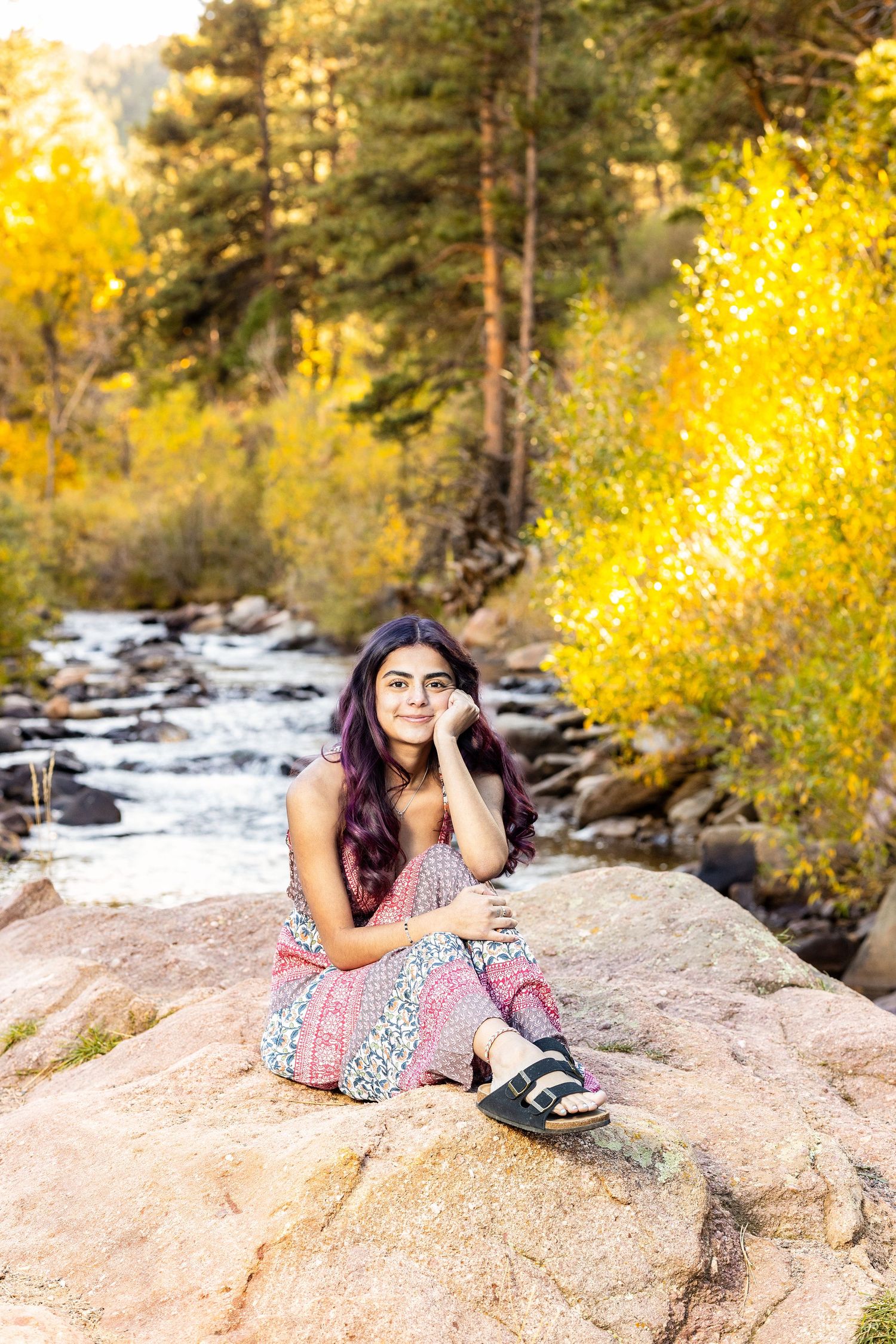 Young woman in floral outfit sitting on rocks by a mountain stream surrounded by golden autumn foliage.