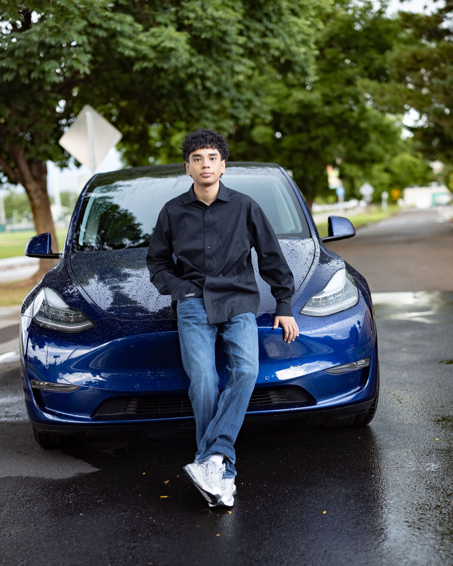 Young man in black shirt and jeans leaning against a blue Tesla on a wet street surrounded by green trees.