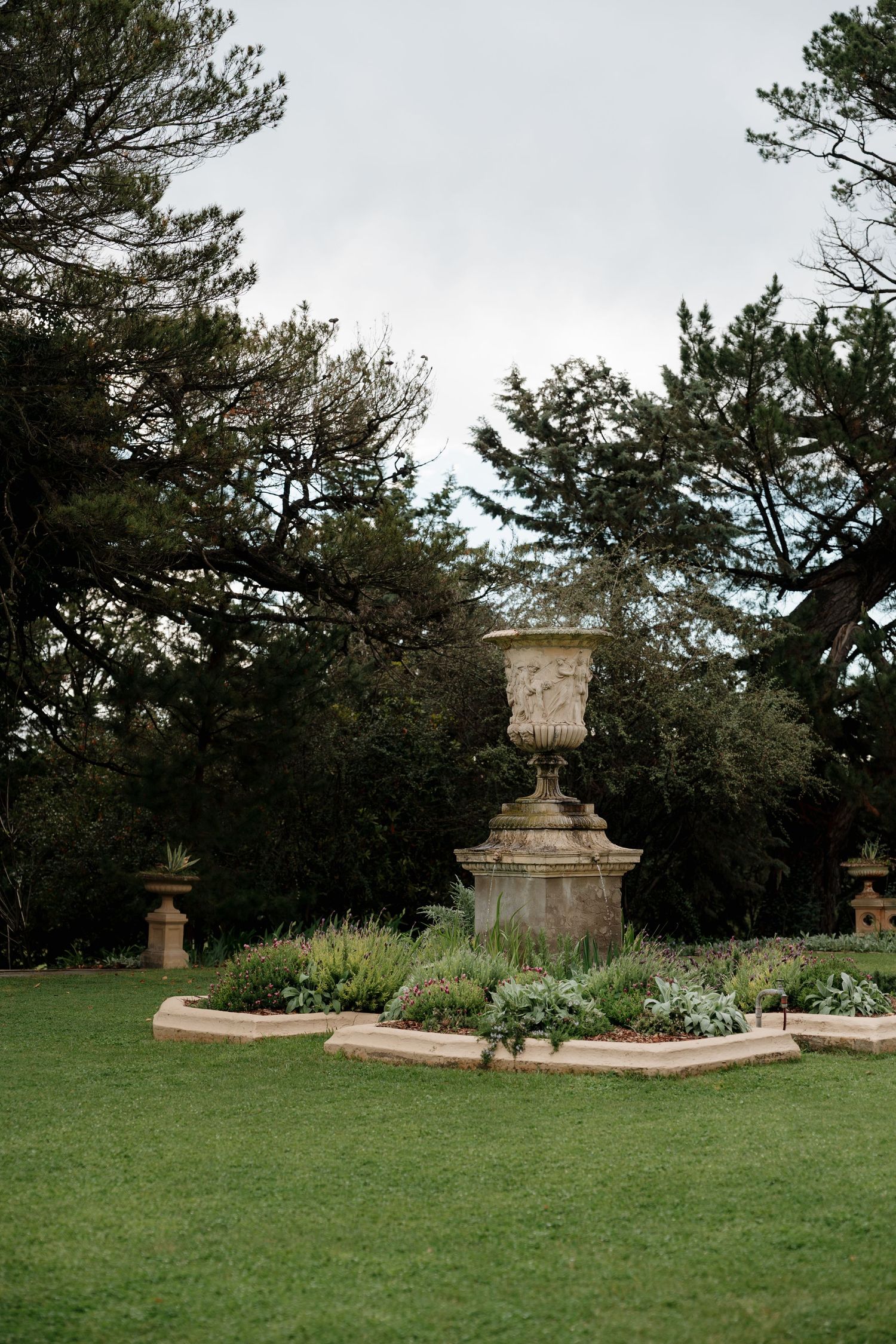 Stone urn garden ornament on pedestal surrounded by lush green plants and tall trees in a formal garden setting.