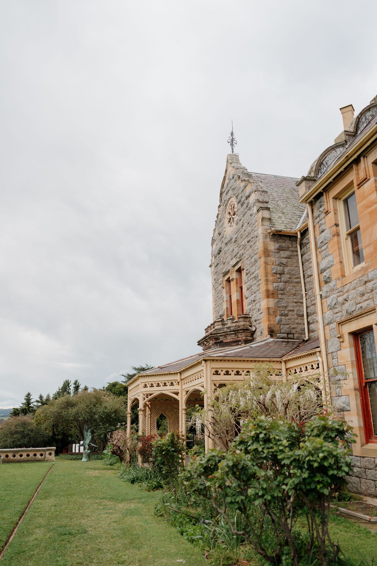 Historic stone mansion with Victorian verandah, ornate wooden trim, and Gothic tower under overcast skies.