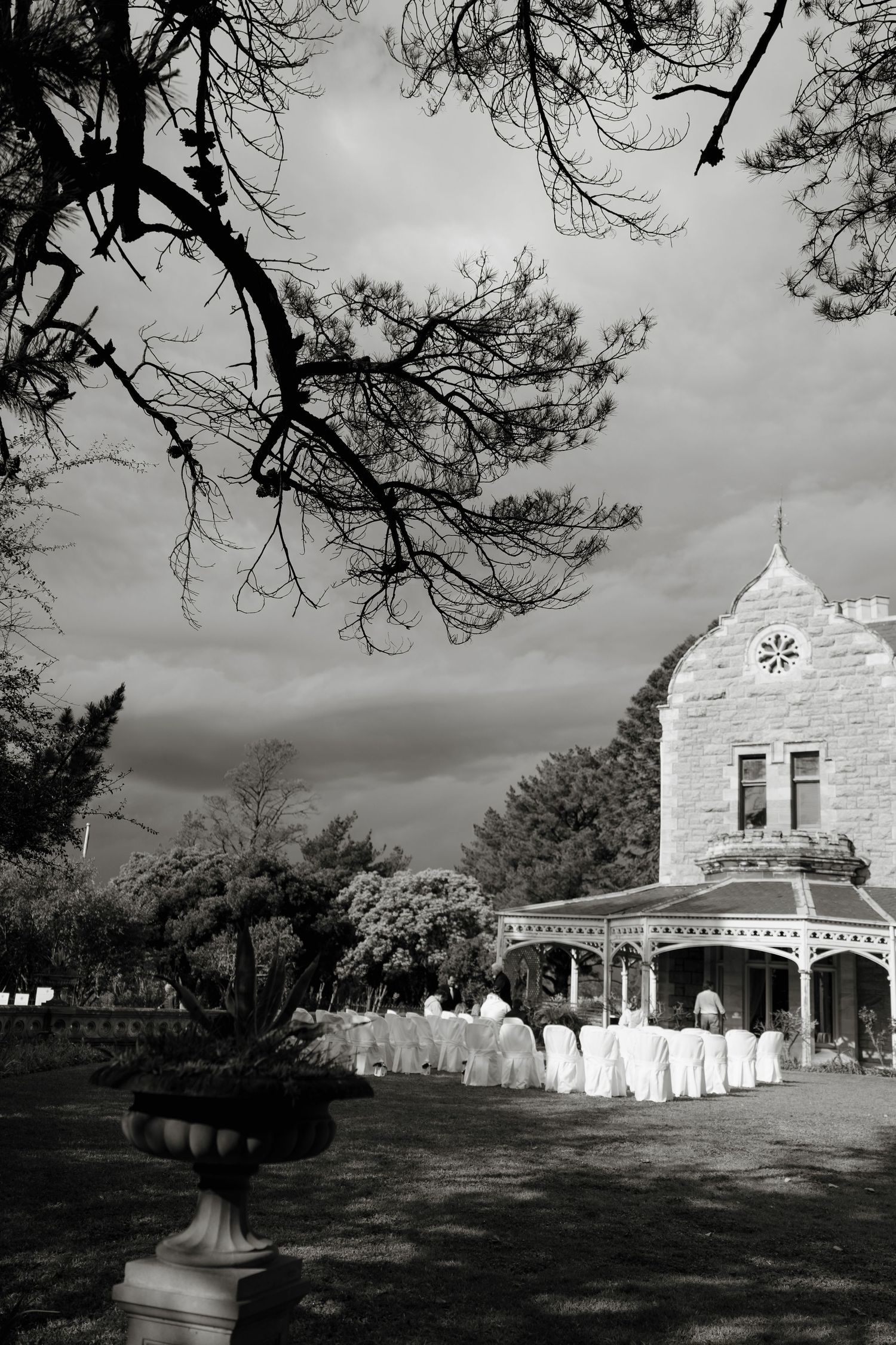 Black and white photo of a historic stone mansion with white chair-lined outdoor wedding setup under dramatic cloudy skies.