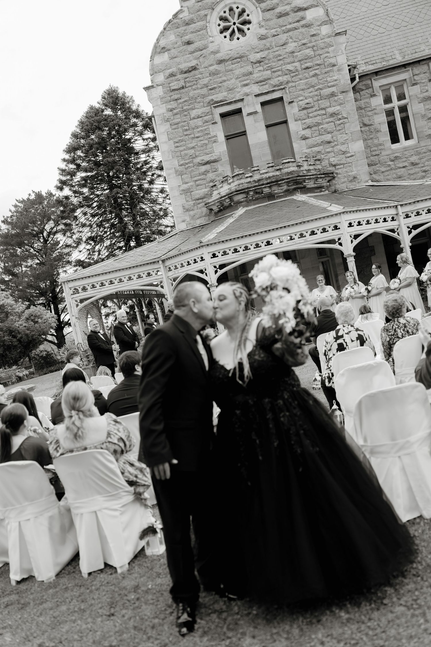 Couple shares first kiss in black wedding attire outside a stone castle venue with seated guests in background.