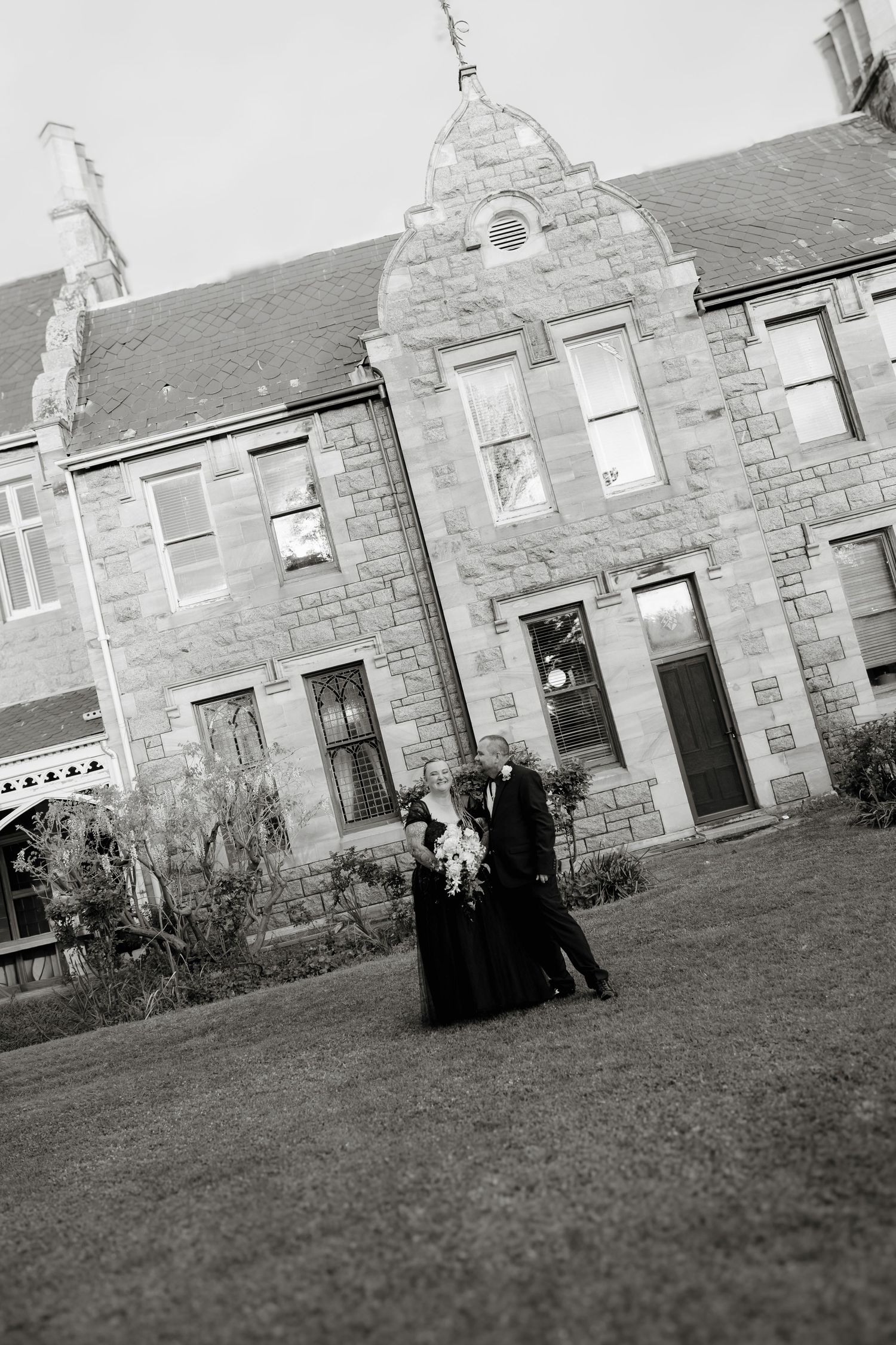Black and white photo of a couple in formal attire posing in front of a historic stone building on a green lawn.