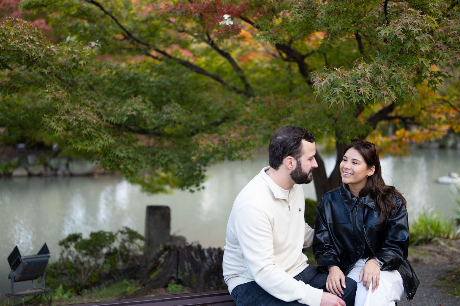 Smiling couple sitting close together on a wooden bench beside a tranquil pond, surrounded by colorful autumn maple leaves.